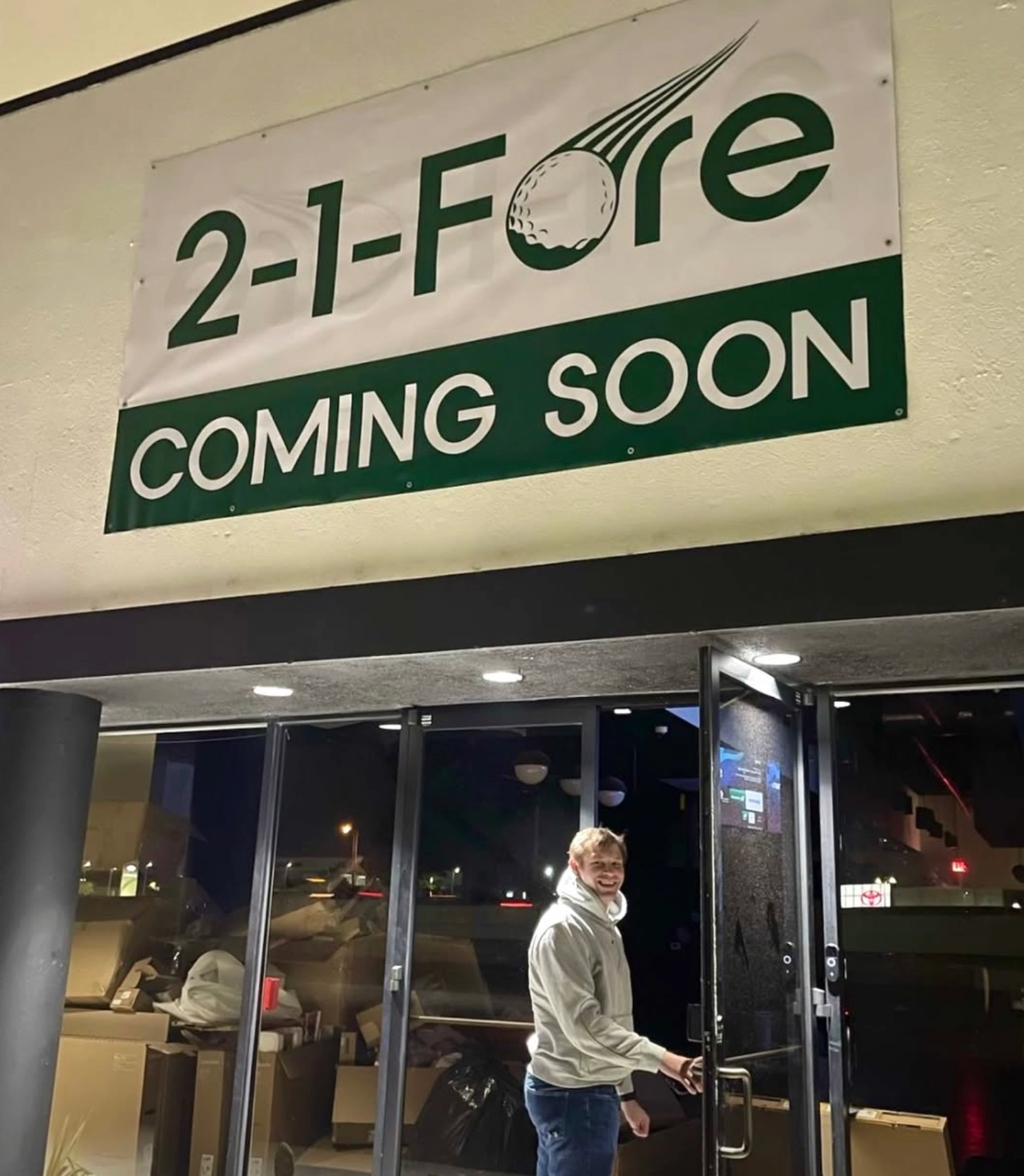 Peter Weldon holding open the door of 2-1-Fore Golf before it opened, under a banner reading 2-1-Fore Coming Soon, with packing boxes stacked inside.