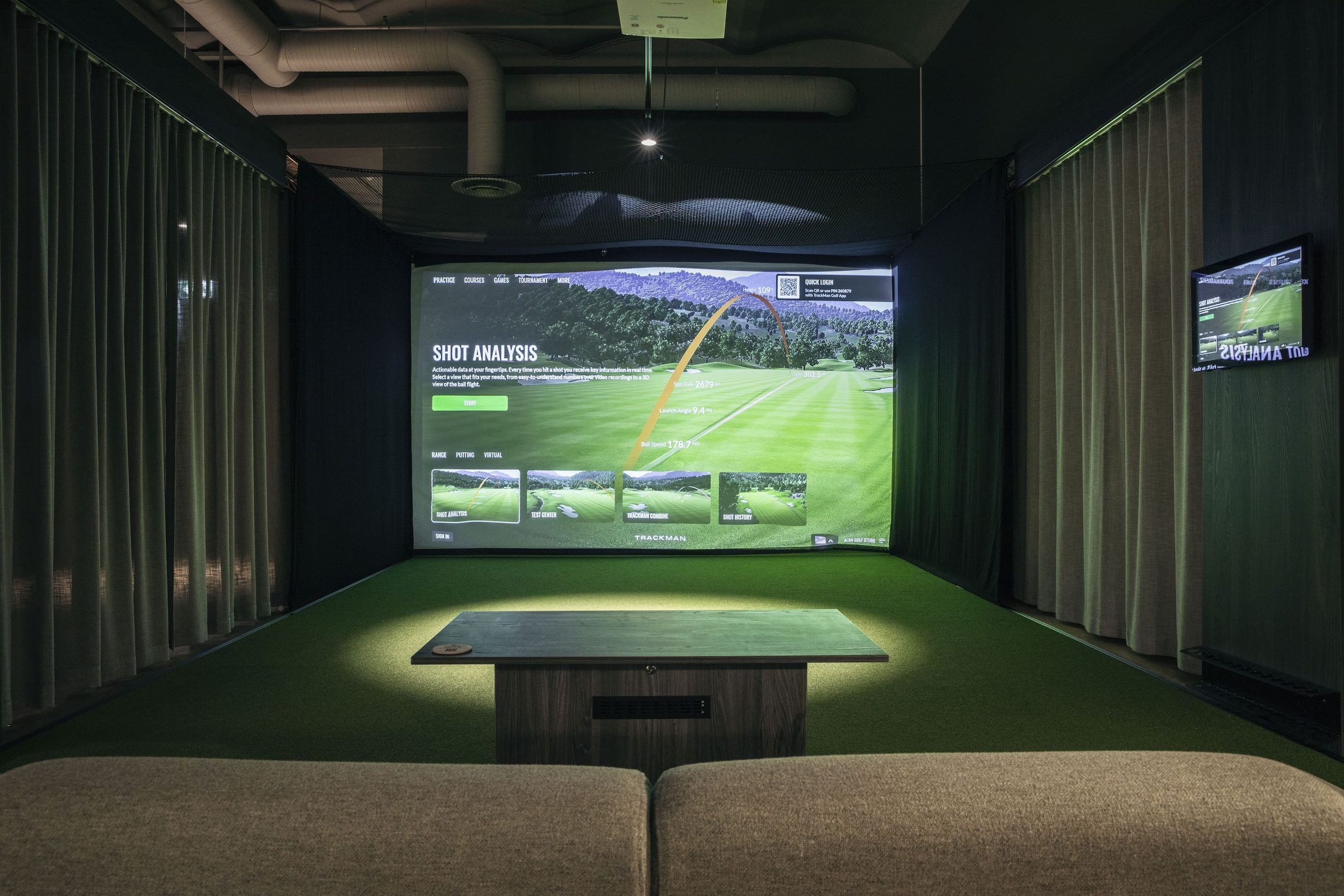 A dark simulator room with a course on a wide screen, a hitting mat and a low lounge table and sofa in front.