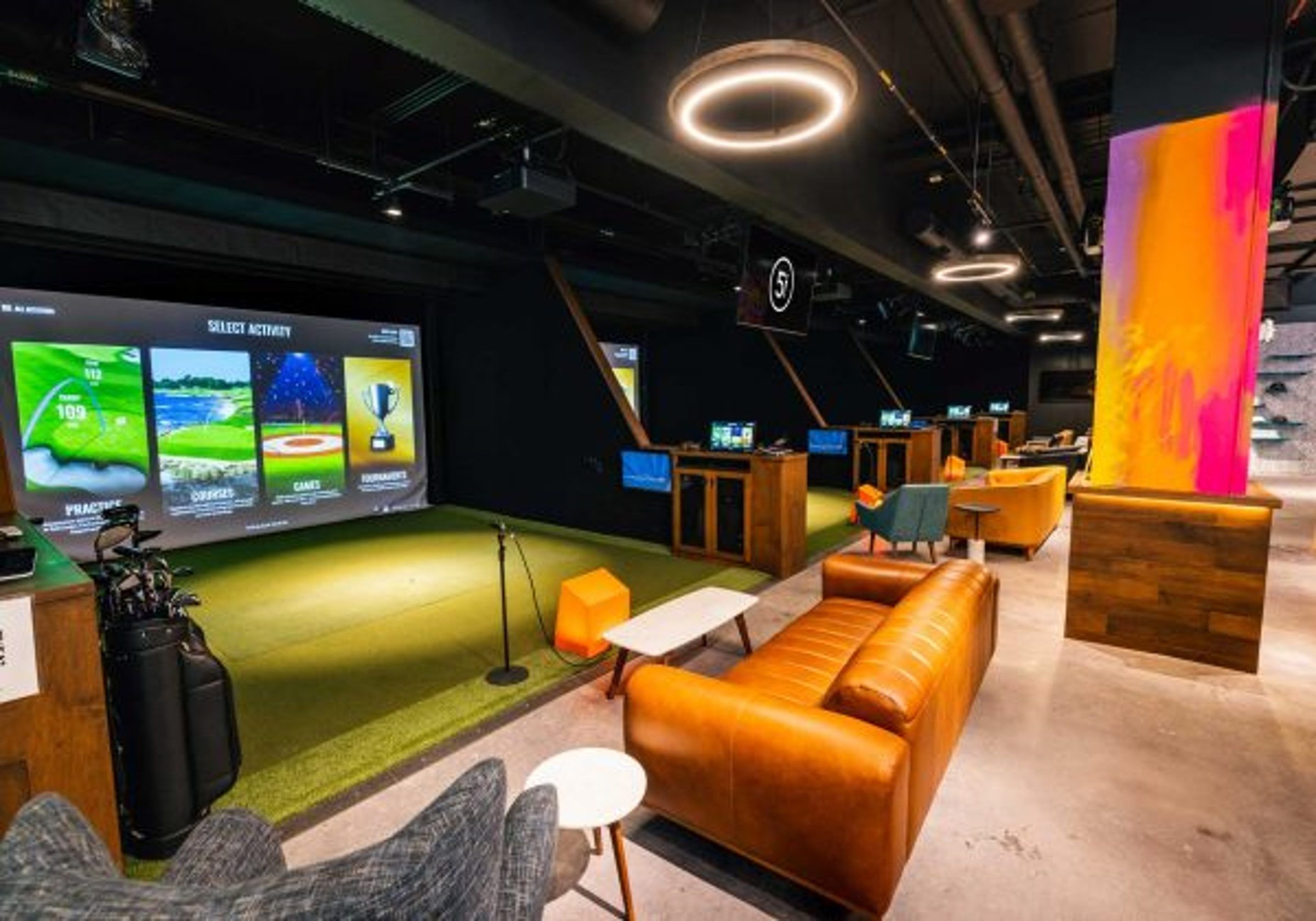 A simulator bay with a green hitting mat and fairway screen, an orange sofa and a bright mural beside it.