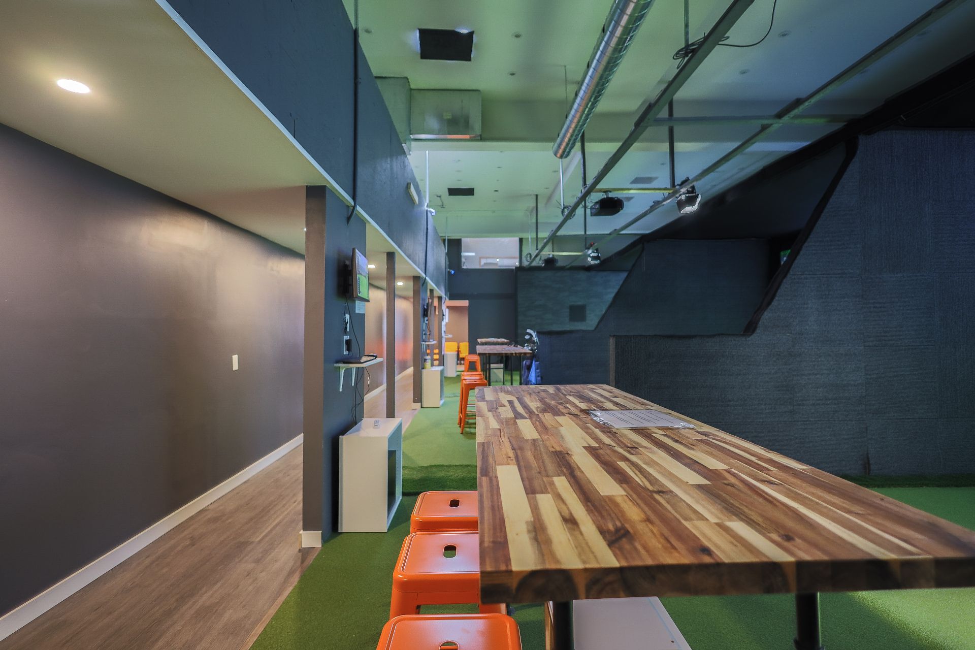 A long butcher-block communal table with orange stools running beside a strip of green turf, under exposed ceiling services.