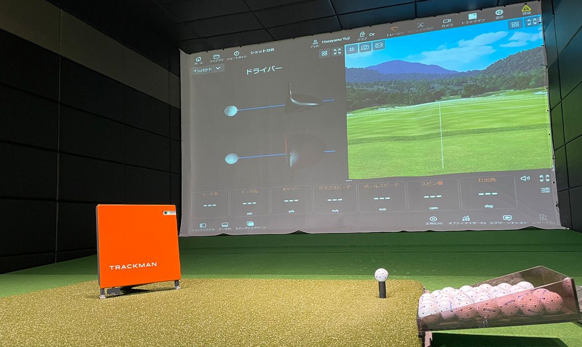 A simulator bay with a fairway on the screen, a hitting mat and a tray of balls on green turf.