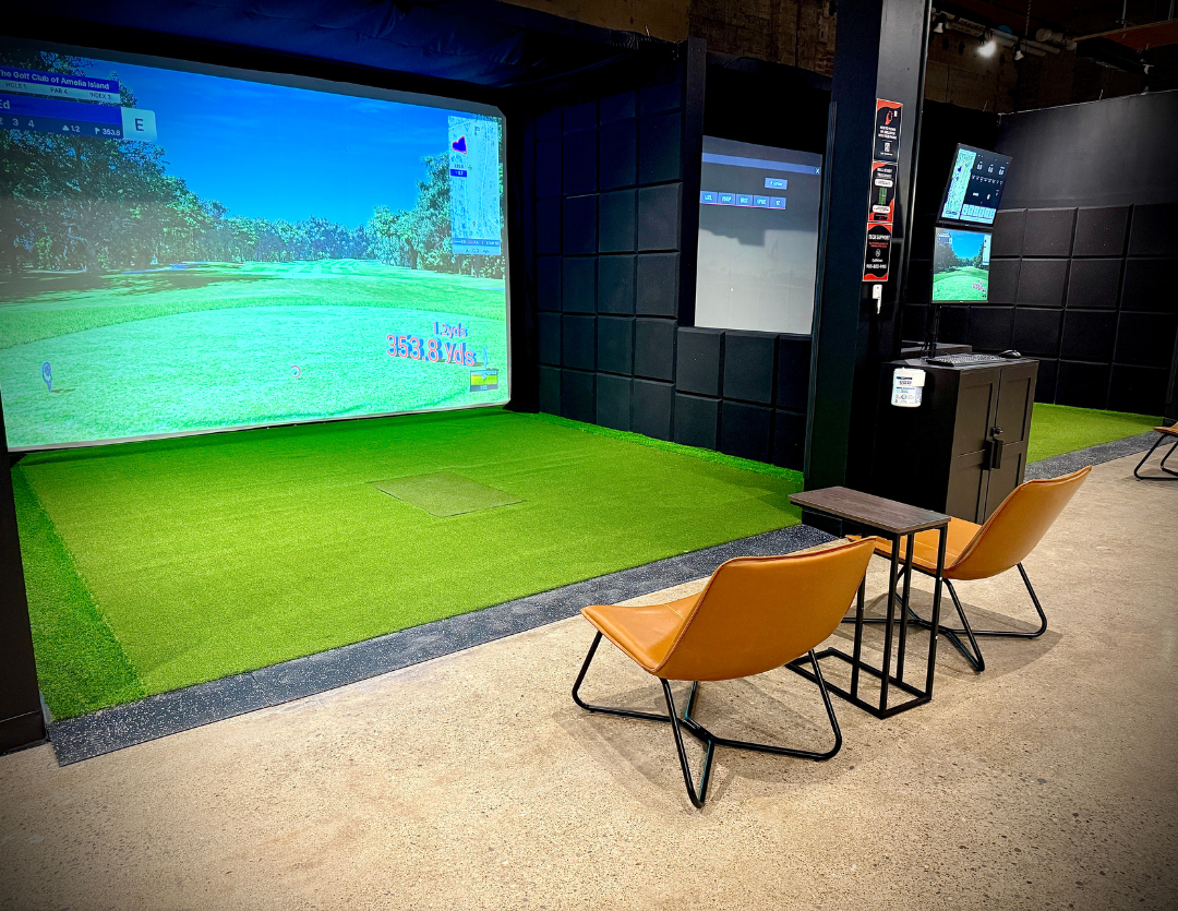 A hitting bay with a coastal course on the screen, green turf underfoot, and two orange chairs beside a small table.