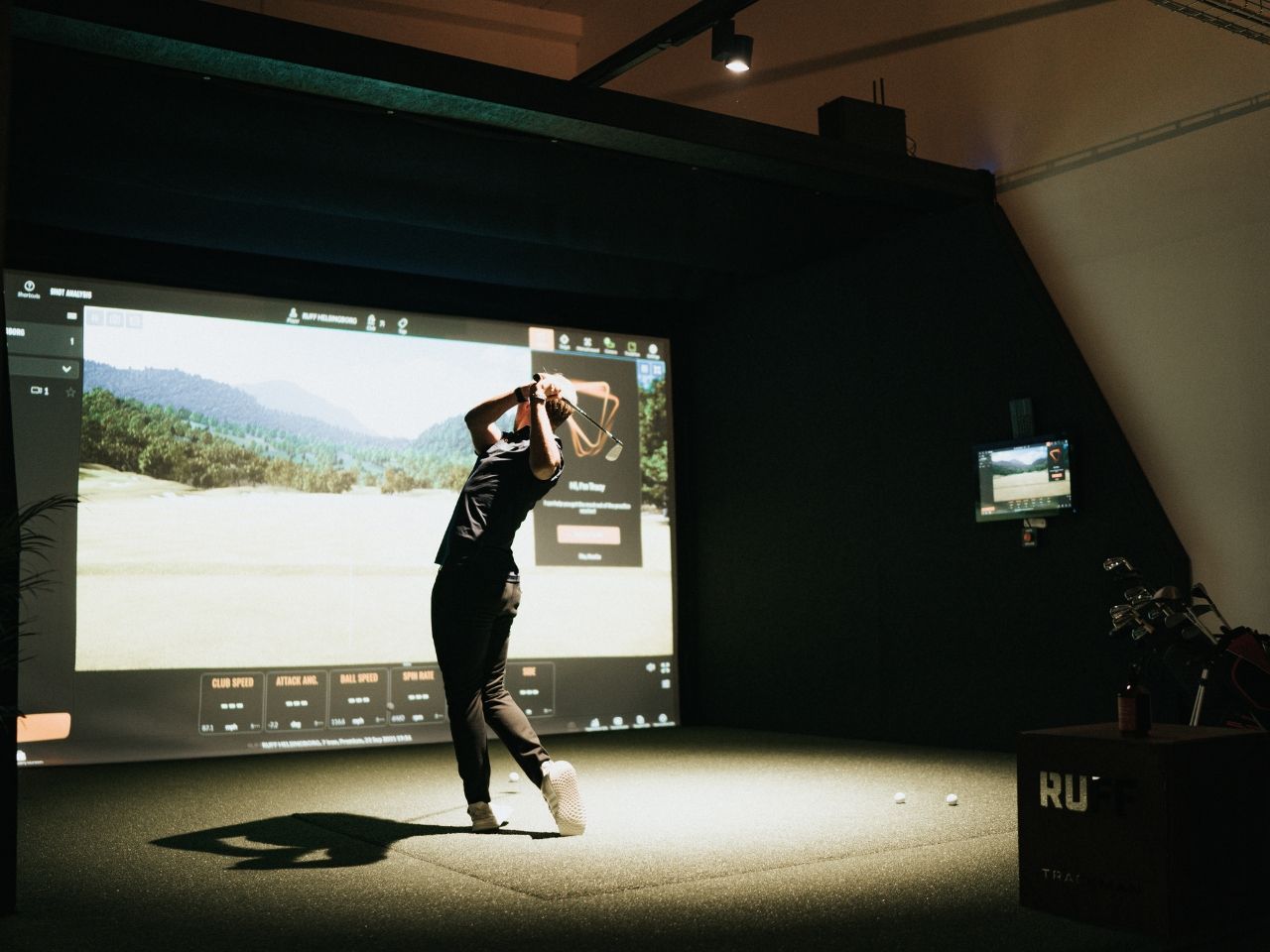 A golfer finishing a swing in a dim simulator bay, with a course on the screen ahead.