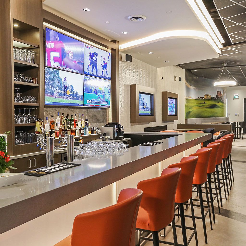 A long white bar counter with orange stools and bottles behind, screens showing golf above.